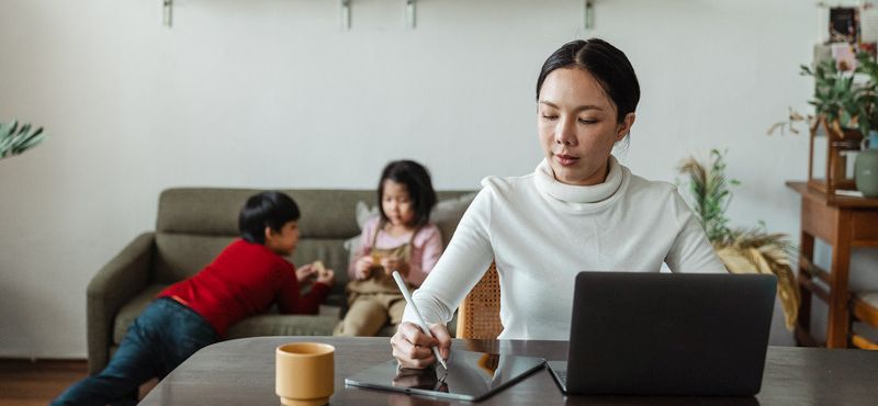 Focused young Asian mother working remotely while children playing on sofa